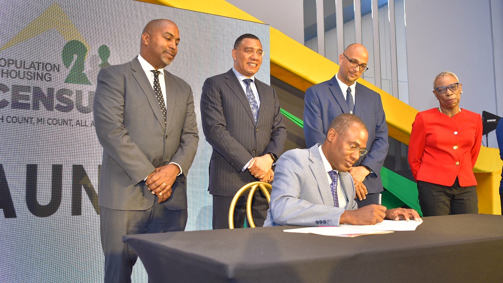 Prime Minister Andrew Holness (second left, standing), observes as Minister of Finance and the Public Service, Dr Nigel Clarke, signs the regulations giving effect to the execution of the 2022 Population and Housing Census, launched at the AC Marriott Kingston Hotel on August 10. Others are (from left) Member of Parliament, St Andrew South Eastern, Julian Robinson (left); Chairman, Professor David Tennant; and Statistical Institute of Jamaica Director General, Carol Coy. (Photo: JIS)