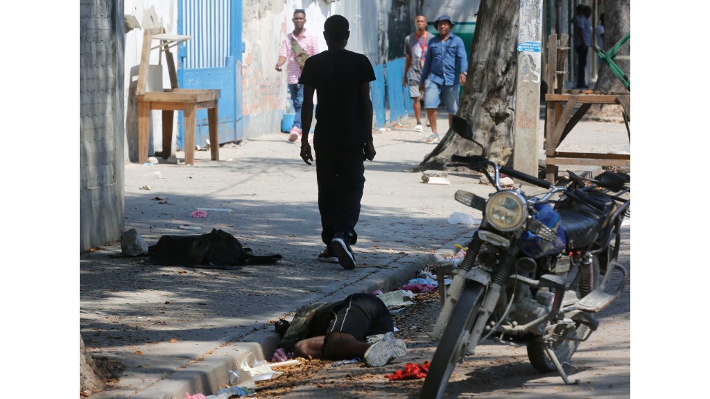 A lifeless body lies against the curb as pedestrians walk past in Port-au-Prince, Haiti, Monday, March 11, 2024. AP Photo:Odelyn Joseph