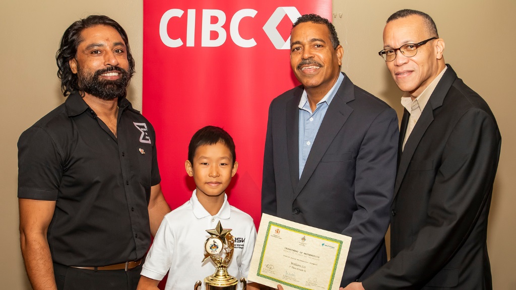 Head of Math Dept (UWI) and Organiser of the Mathematical Olympiad Dr Mahesha Narayama (left) stands alongside 9 year-old old William Lei, winner of the grade six Junior Mathematical Olympiad Competition. The young prodigy still in the 4th grade stands proudly with his championship trophy as CIBC Managing Director Nigel Holness (2nd right) and Douglas Cupidon Director of Credit, Sovereigns,and Financial Institutions from CIBC hold his 1st place certificate.